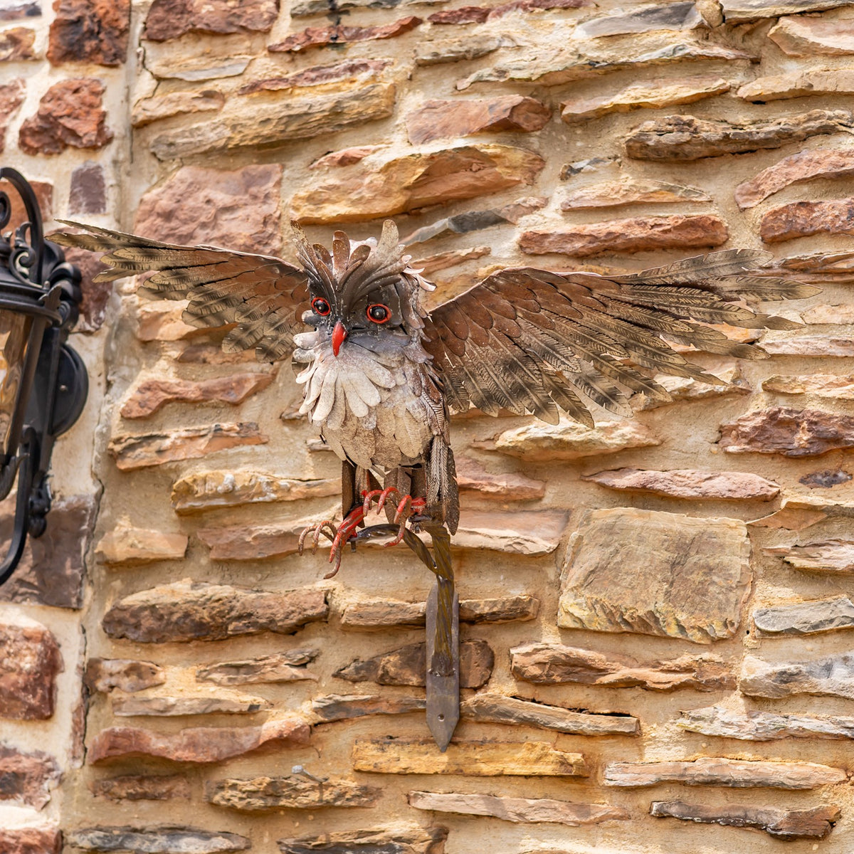 Front view of a metal owl with broad outstretched wings and red feet on a branch by a stone wall.