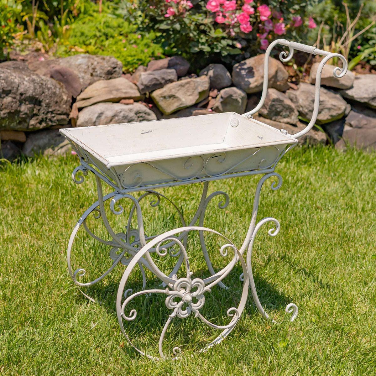 Empty antique-white flower cart on the lawn, with a shallow tray, curled handle and decorative wheels.