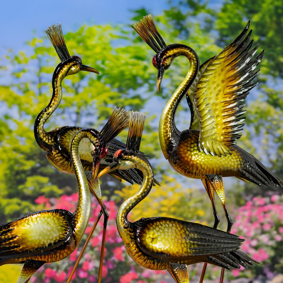 Several metallic heron figurines pose together, including one with a raised wing, among garden flowers.