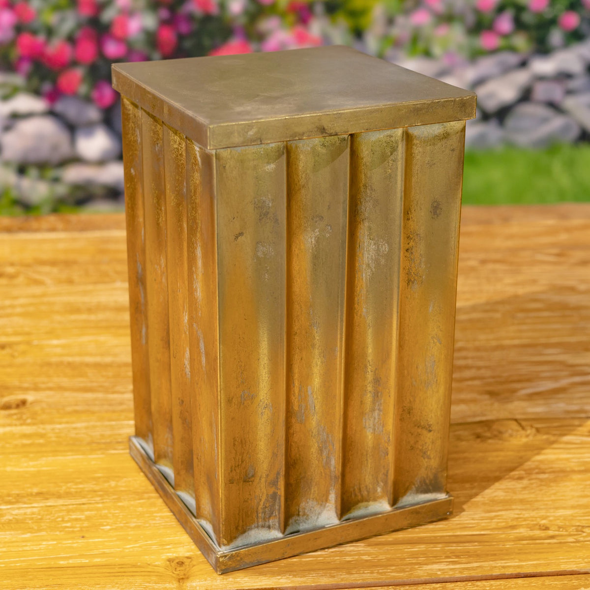 A gold-toned metal pedestal pillar with a square top and vertically fluted sides, photographed outdoors.