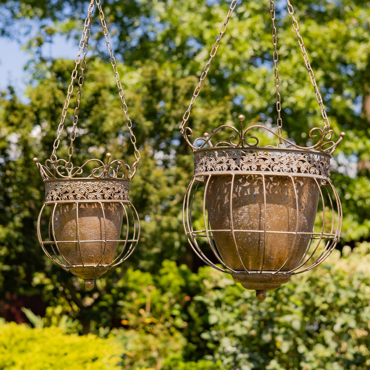 Two ornate metal hanging planters in different sizes suspended against a leafy garden backdrop.