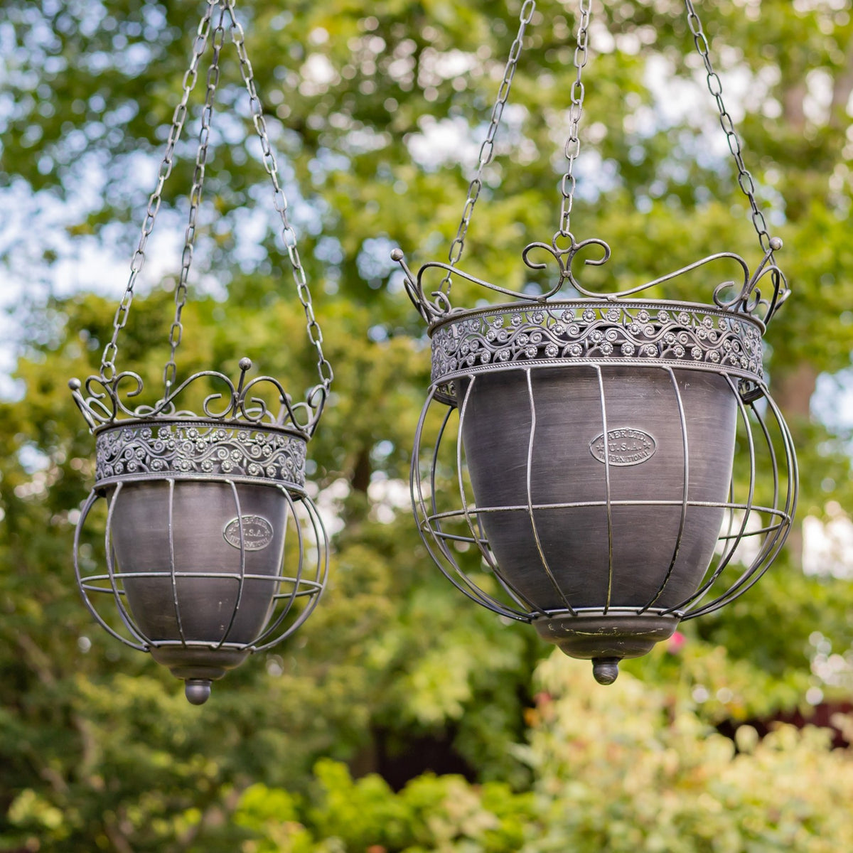 Two empty ornate metal hanging planters shown together against trees, in different sizes.