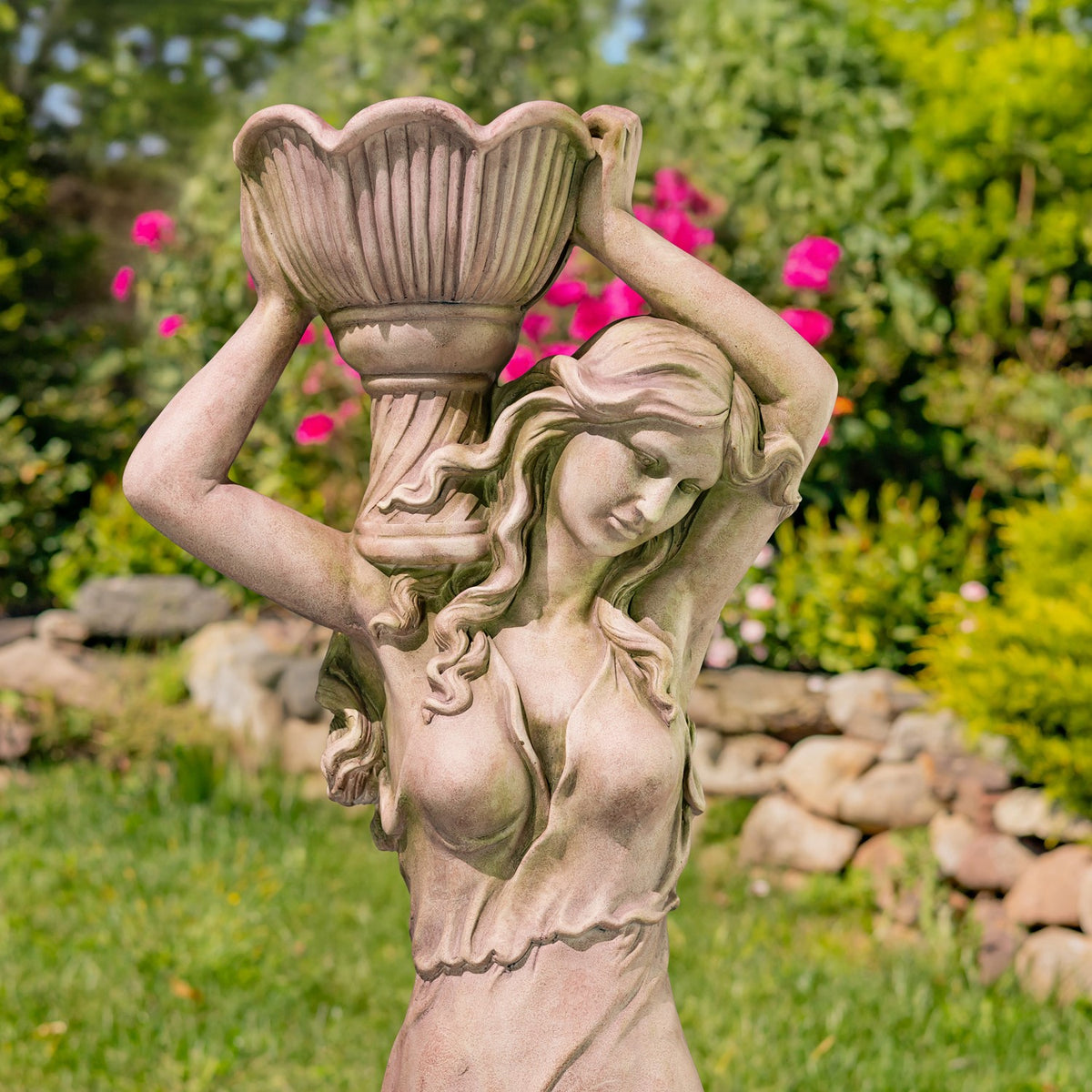 Close view of the statue’s raised arms, flowing hair, and fluted pot against a garden backdrop.