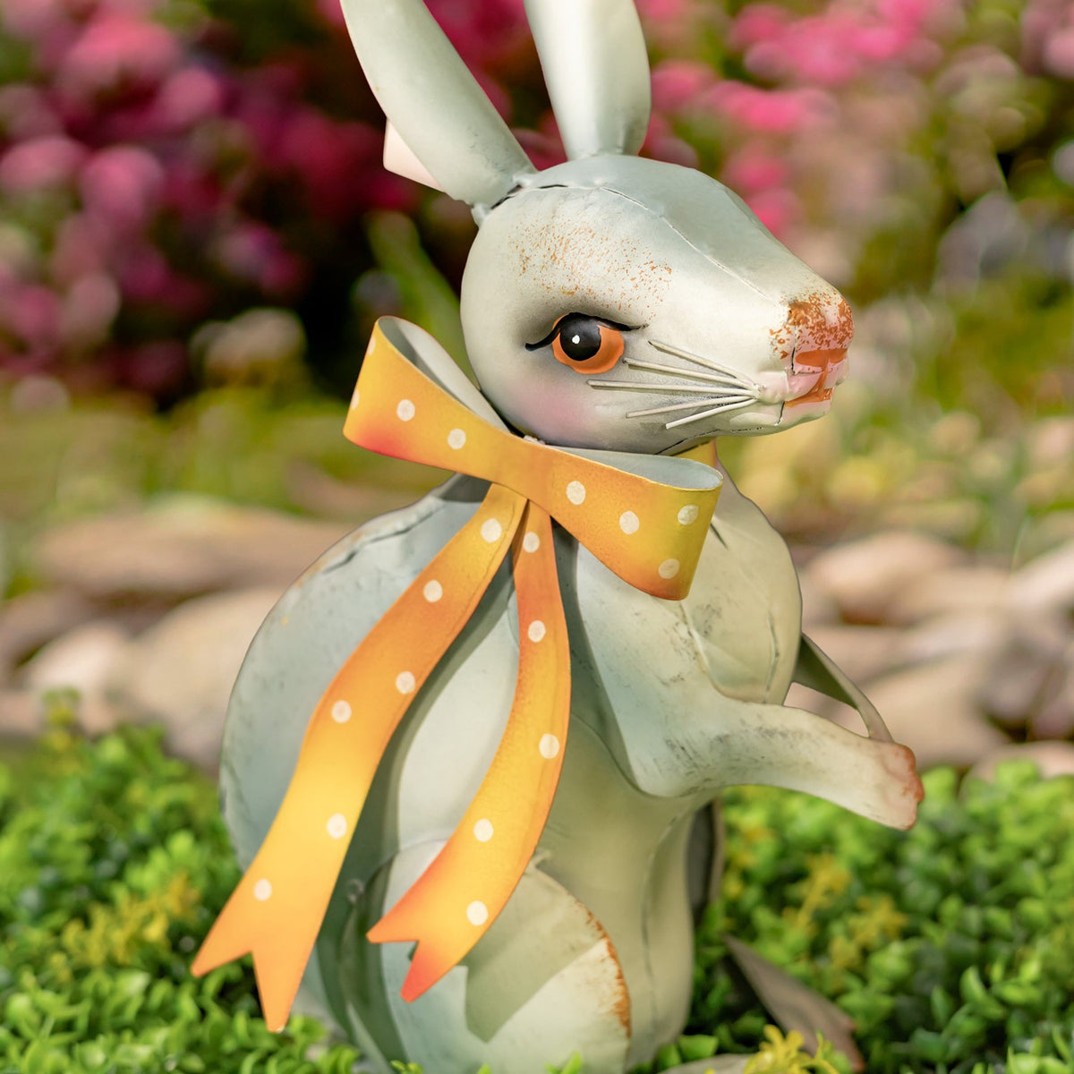 A seated rabbit figurine with a dotted orange ribbon is shown against blurred pink flowers and garden stones.
