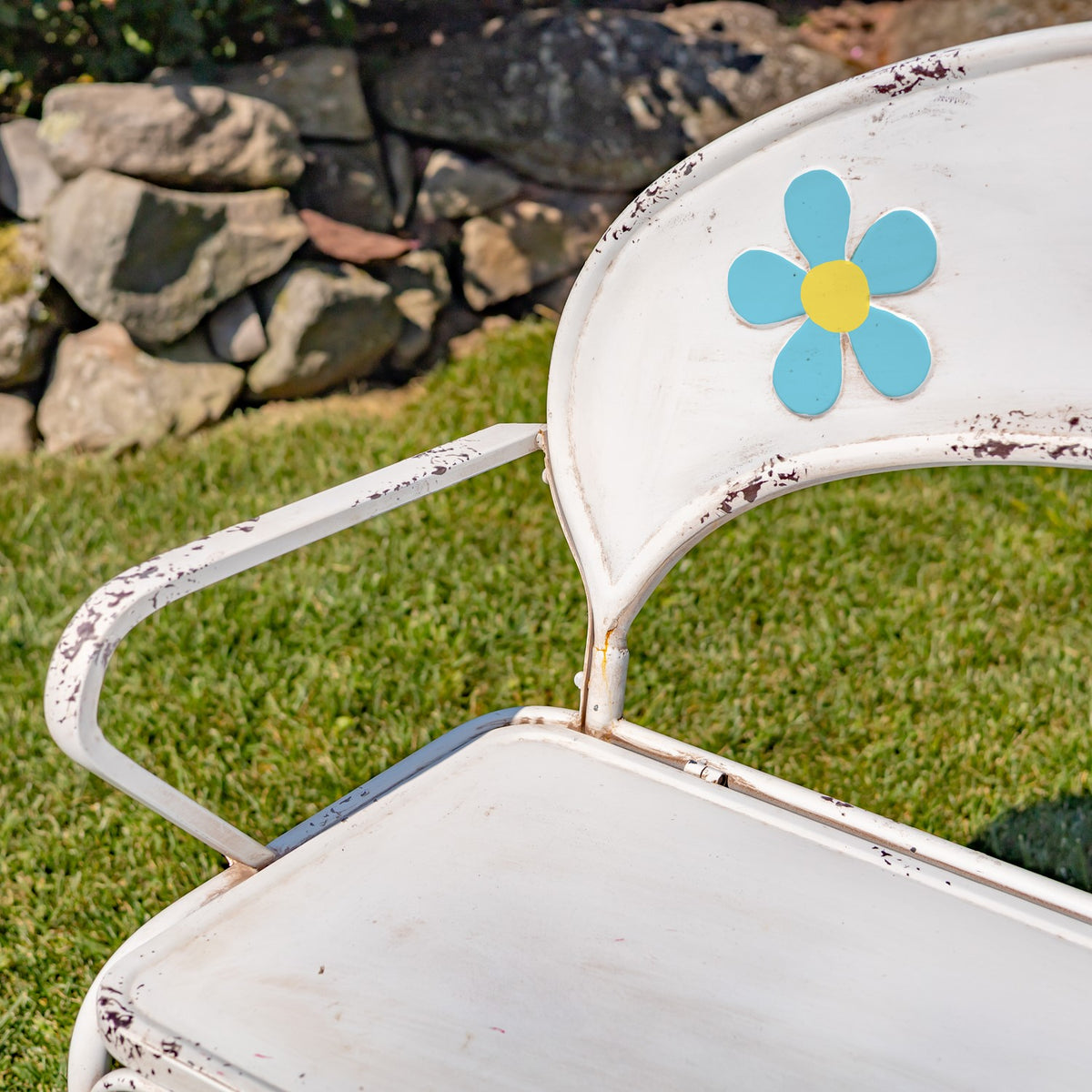 Close-up of a white garden bench with a blue-and-yellow flower on the backrest and a weathered finish.