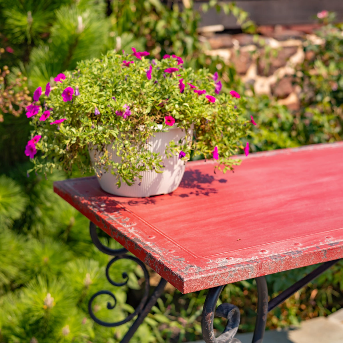 Red rectangular tabletop with a white flower planter, supported by ornate black iron legs in a garden.
