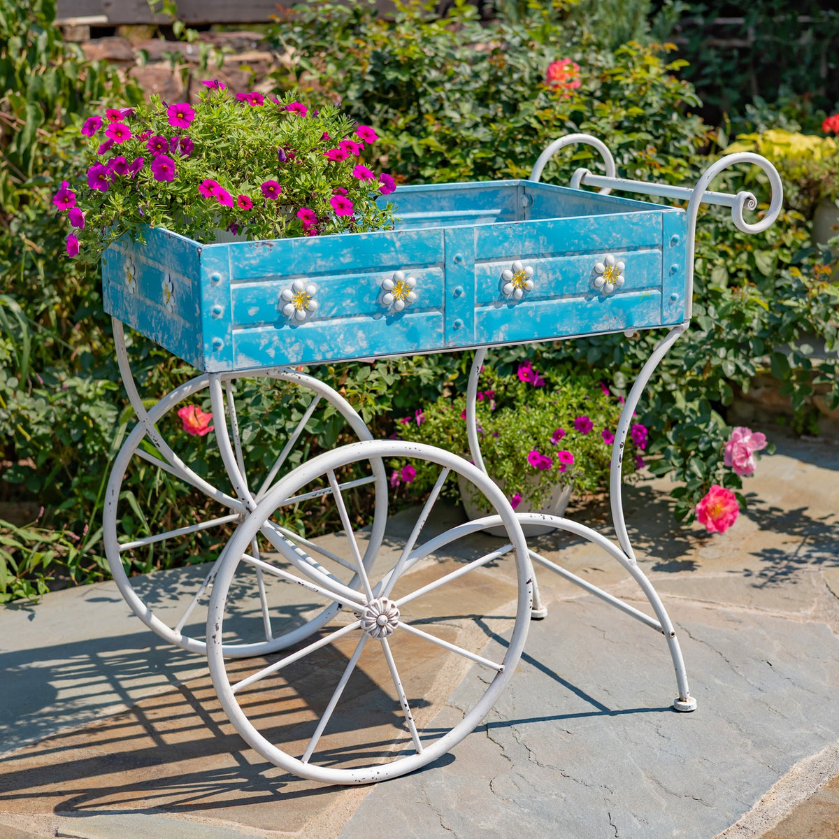 Blue flower cart filled with pink flowers, with decorative panels, large spoked wheels, and a curled handle.