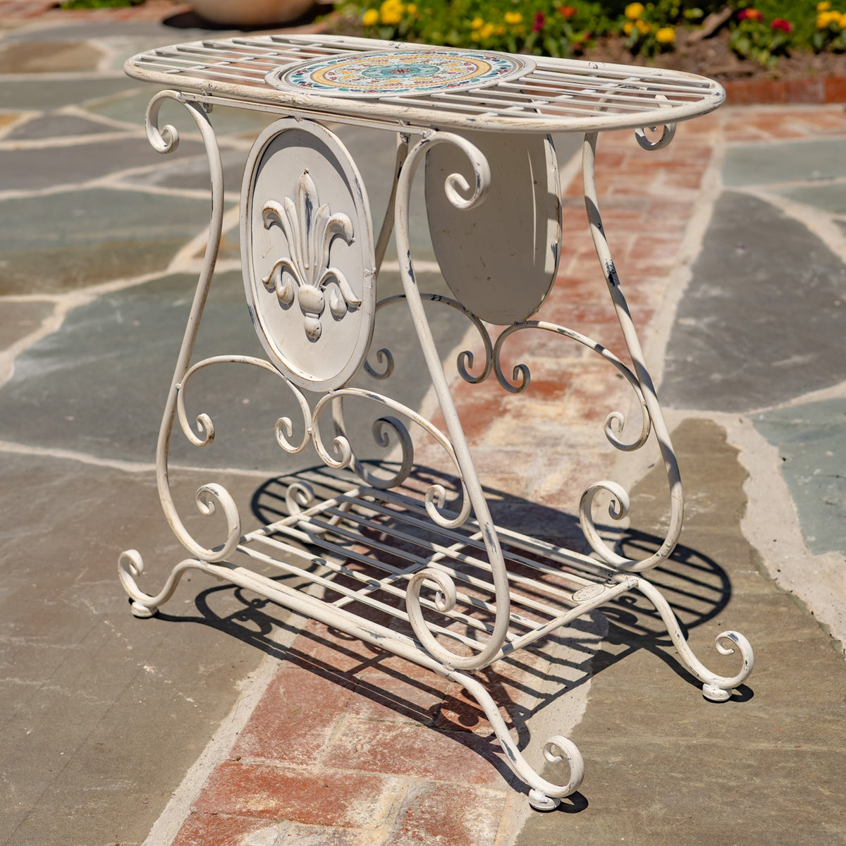 Close view of a cream plant stand with a colorful mosaic inset, fleur-de-lis panel, and curled metalwork.