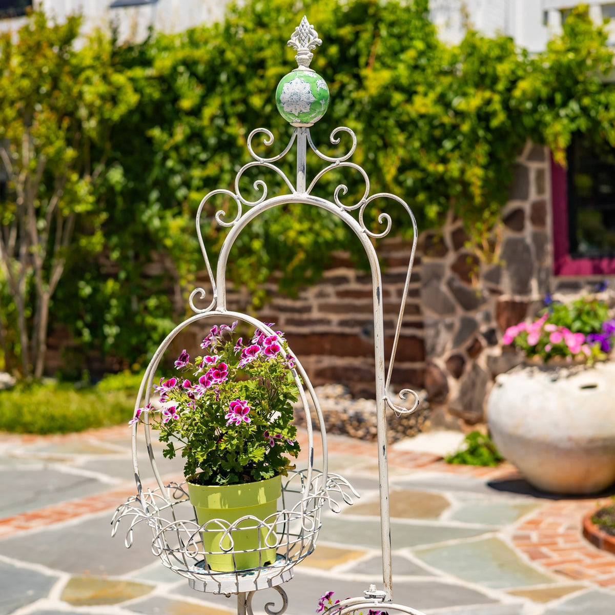 Closer view of the upper wire basket holding a green pot of pink flowers beneath scrollwork.