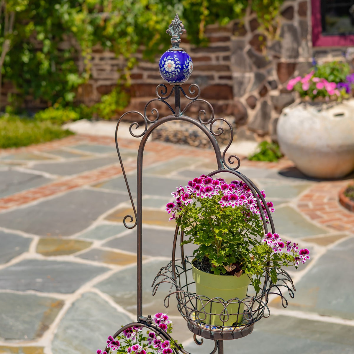 Closer view of the stand’s blue floral finial, curled metal arch, and upper basket holding pink flowers.
