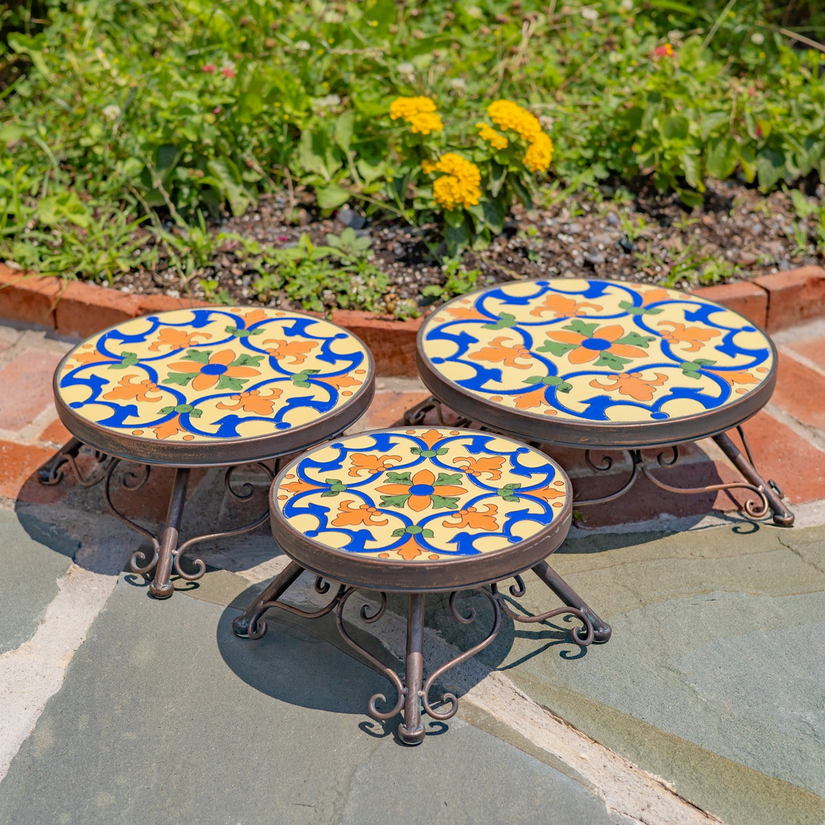 Three empty round plant stands with colorful floral mosaic tops arranged at different heights outdoors.