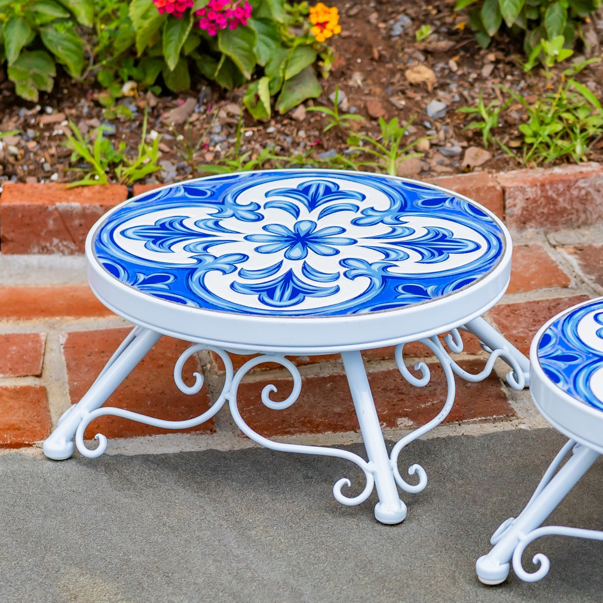 A round stand with a blue floral-patterned top and curled white legs beside a brick garden border.