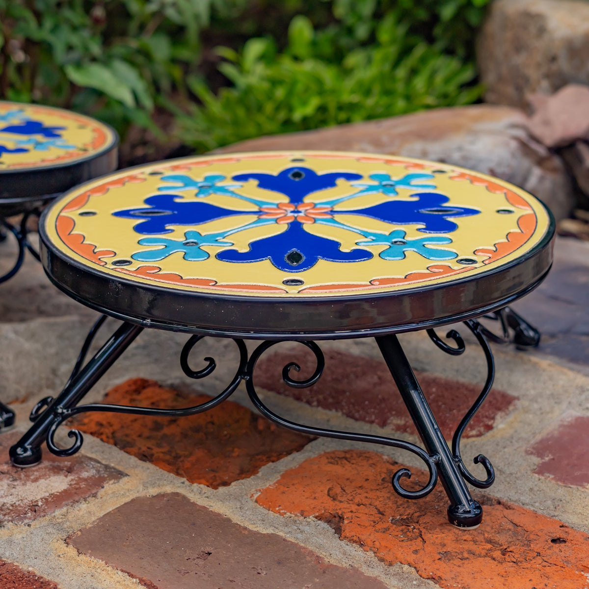 A round stand shows its bright floral mosaic top and curled black metal legs.