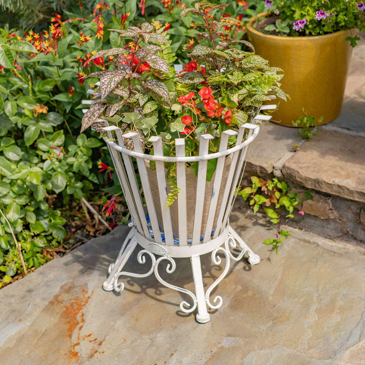 White slatted iron basket stand with ornate feet among flowers and greenery on a stone patio.