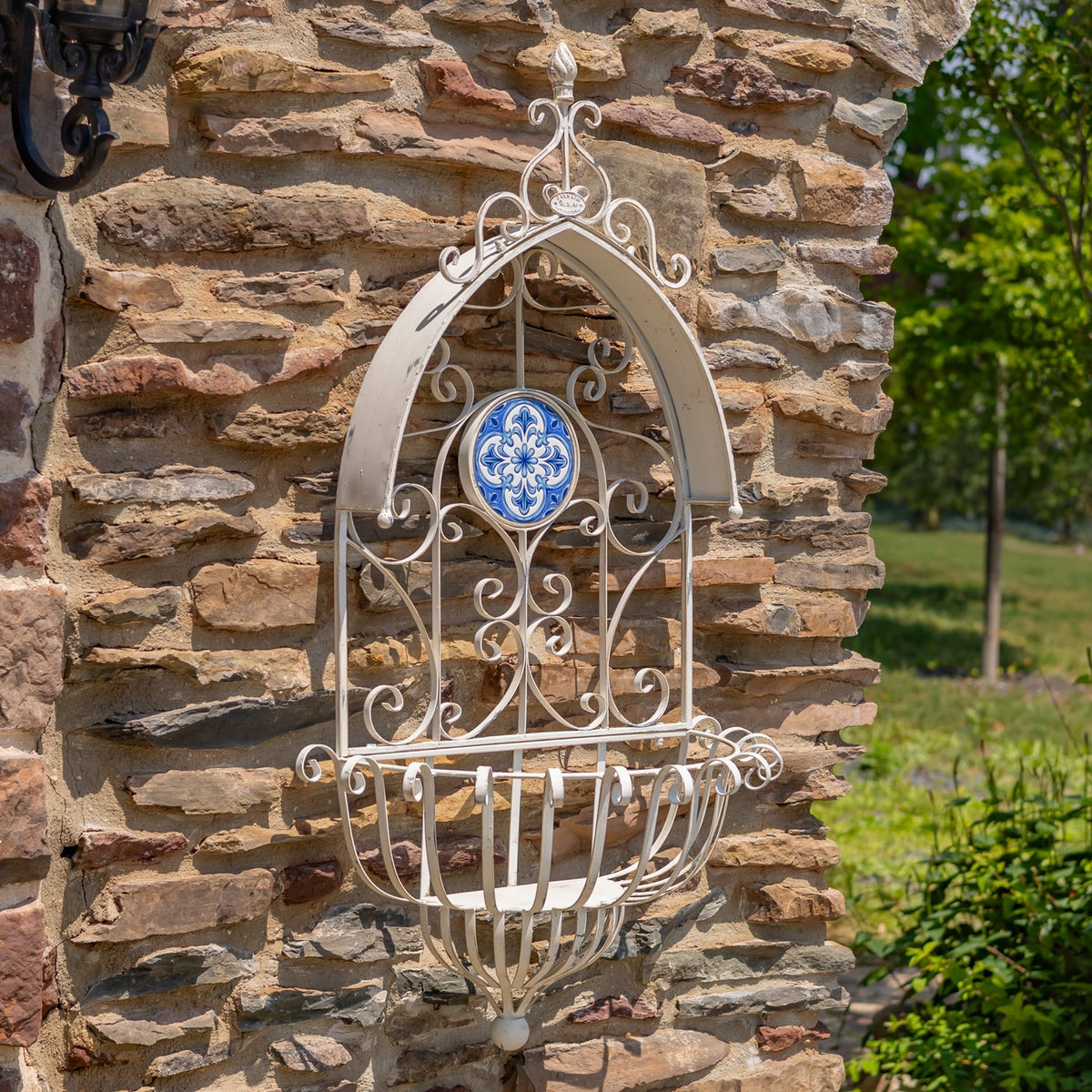 Unfilled white scrollwork planter with a blue patterned tile displayed on a rustic stone wall.