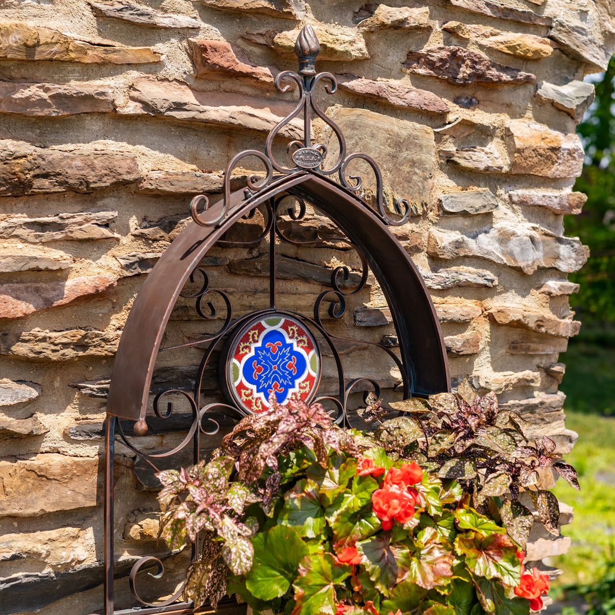 Arched wrought-iron wall planter with a bright mosaic medallion and leafy plants with red blooms in its basket.