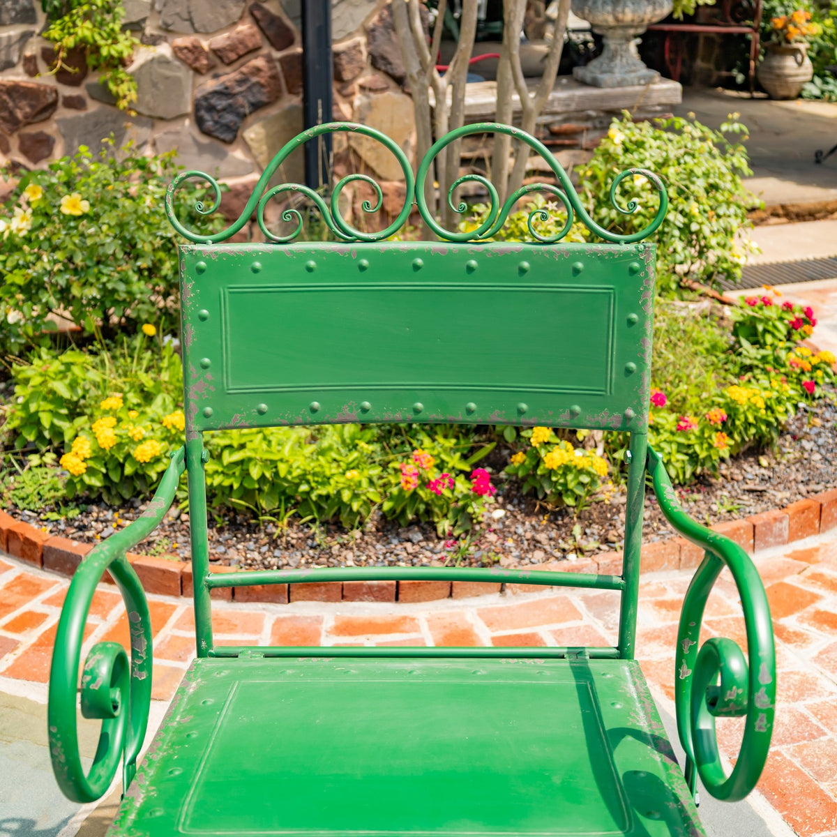 Rear view of a green metal patio chair with a scrollwork back and curved arms in a flower garden.