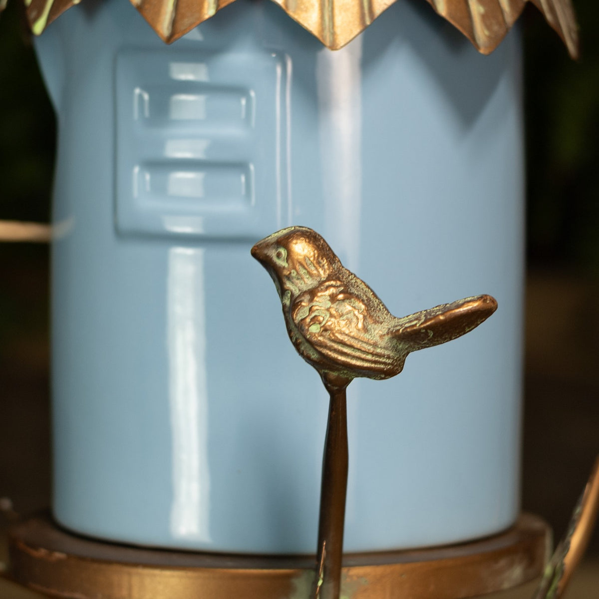 Gold-toned bird ornament on a slender stem in front of the blue birdhouse body.