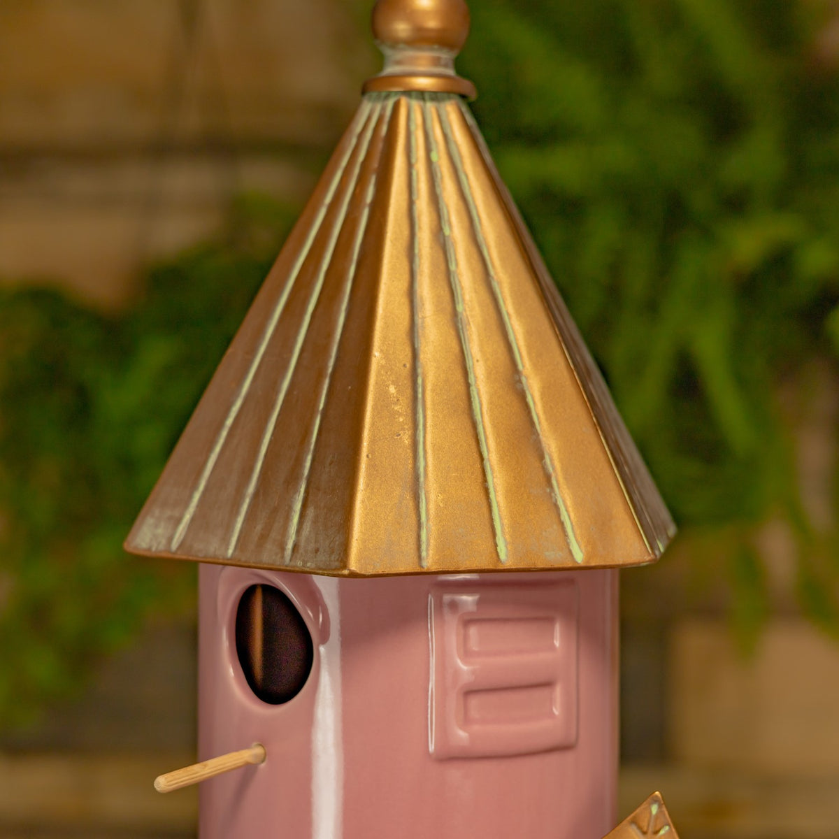 Close view of the birdhouse’s round entrance, short perch, pink body and ribbed gold-toned roof.