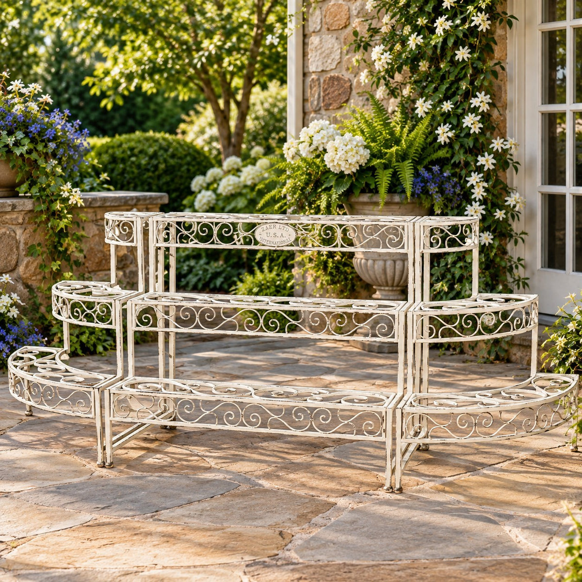 Ornate tiered metal plant stand displayed on a sunny patio beside a stone wall and climbing flowers.