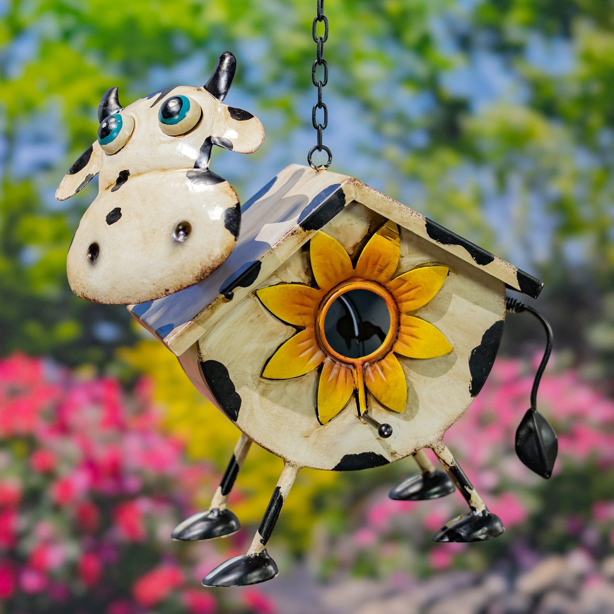 Spotted cream cow-shaped birdhouse with a sunflower entrance and dangling tail, against flowers.