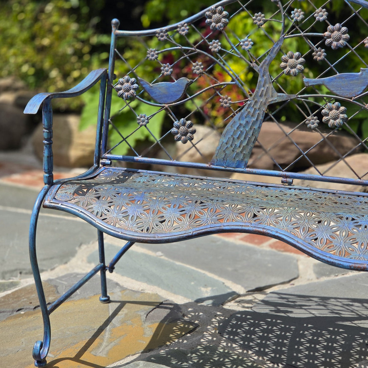 Close view of the bench’s pierced floral seat, curved arm, and bird-and-tree backrest design.