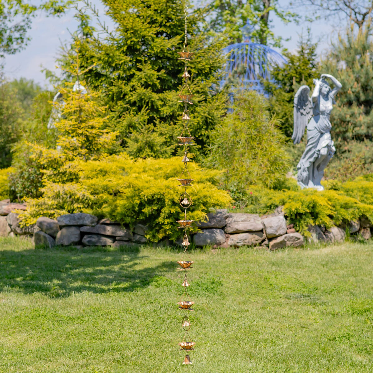 The full rain chain hangs vertically over a lawn, with a stone border, trees, and a garden statue behind it.