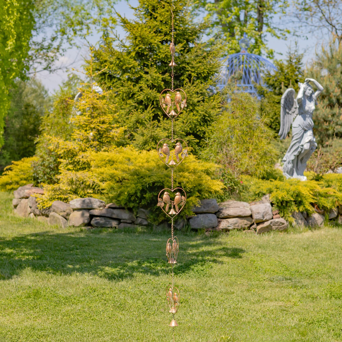 The rain chain hangs nearly to the lawn, showing several bird-and-bell heart ornaments in a landscaped garden.