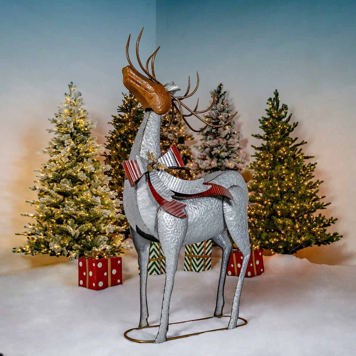 A silver reindeer with brown antlers and a striped bow stands on a metal base before Christmas trees.