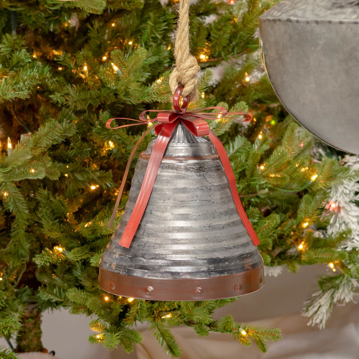 A tapered galvanized bell with a red ribbon and bow hangs against green Christmas tree branches.