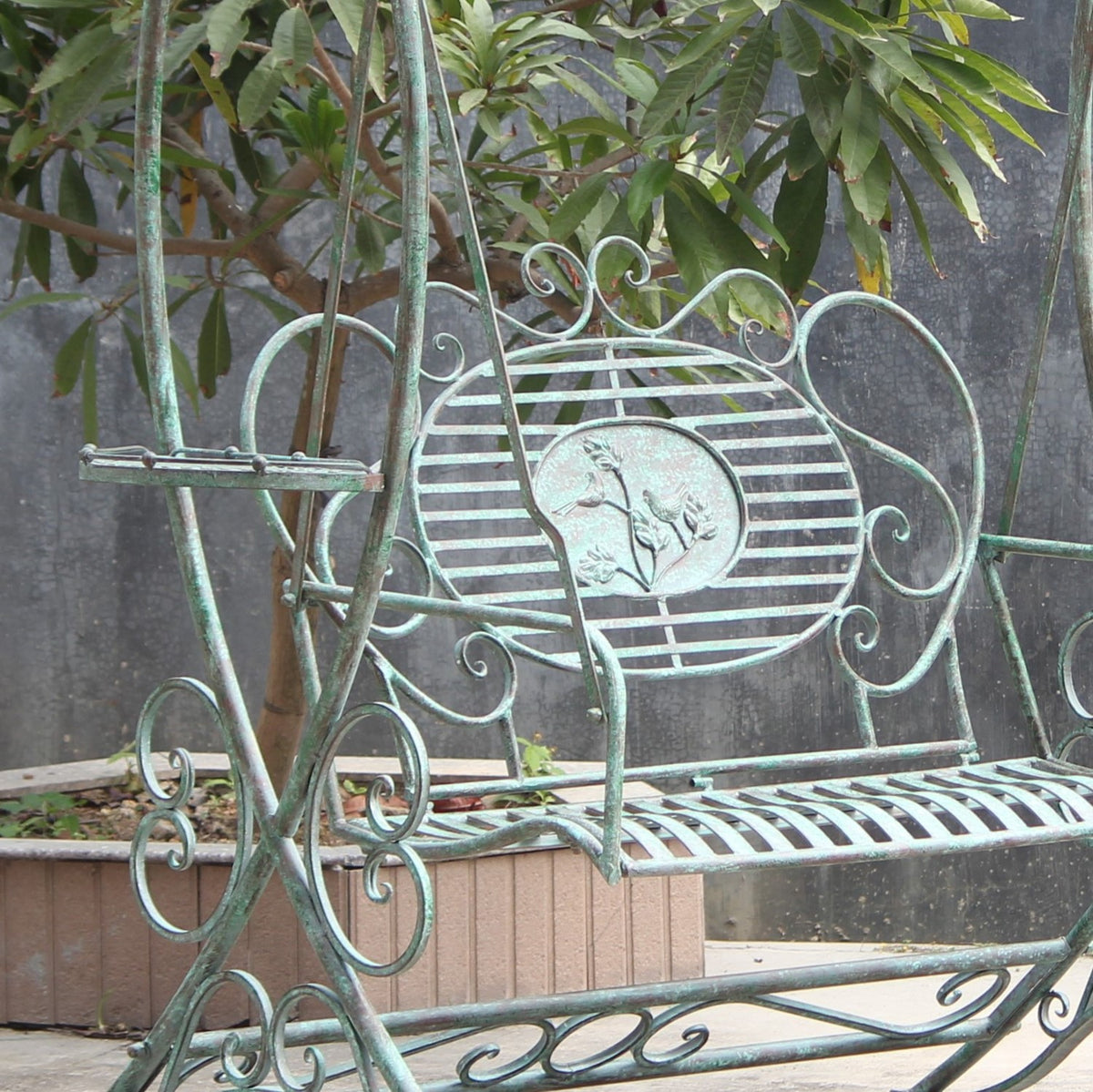 Ornate metal garden swing bench with a slatted seat, decorative backrest, and small side shelves among plants.