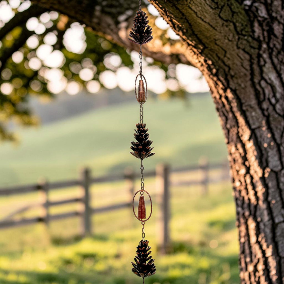 Pinecone-shaped metal ornaments and oval cups hang in a vertical chain beside a tree trunk.