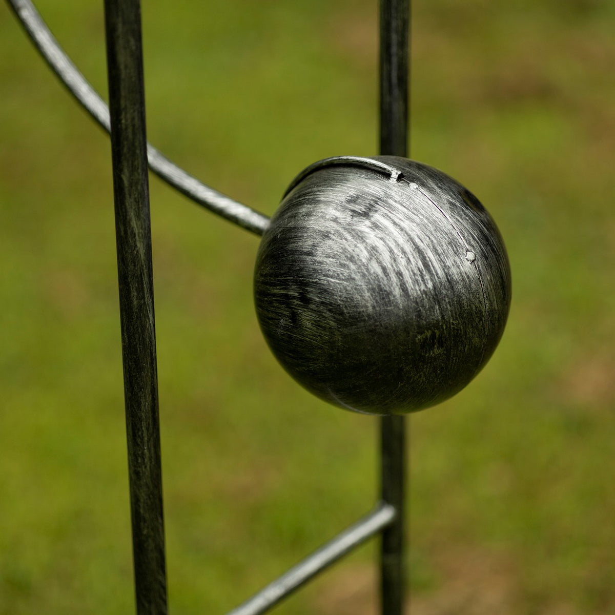 A rounded dark metal ball hangs between the vertical rods of the garden stake.