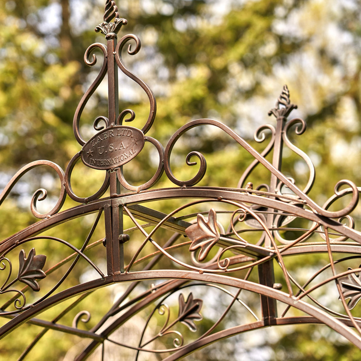 Decorative iron arch crest with curled scrolls and leaf motifs, photographed against a sunlit garden backdrop.