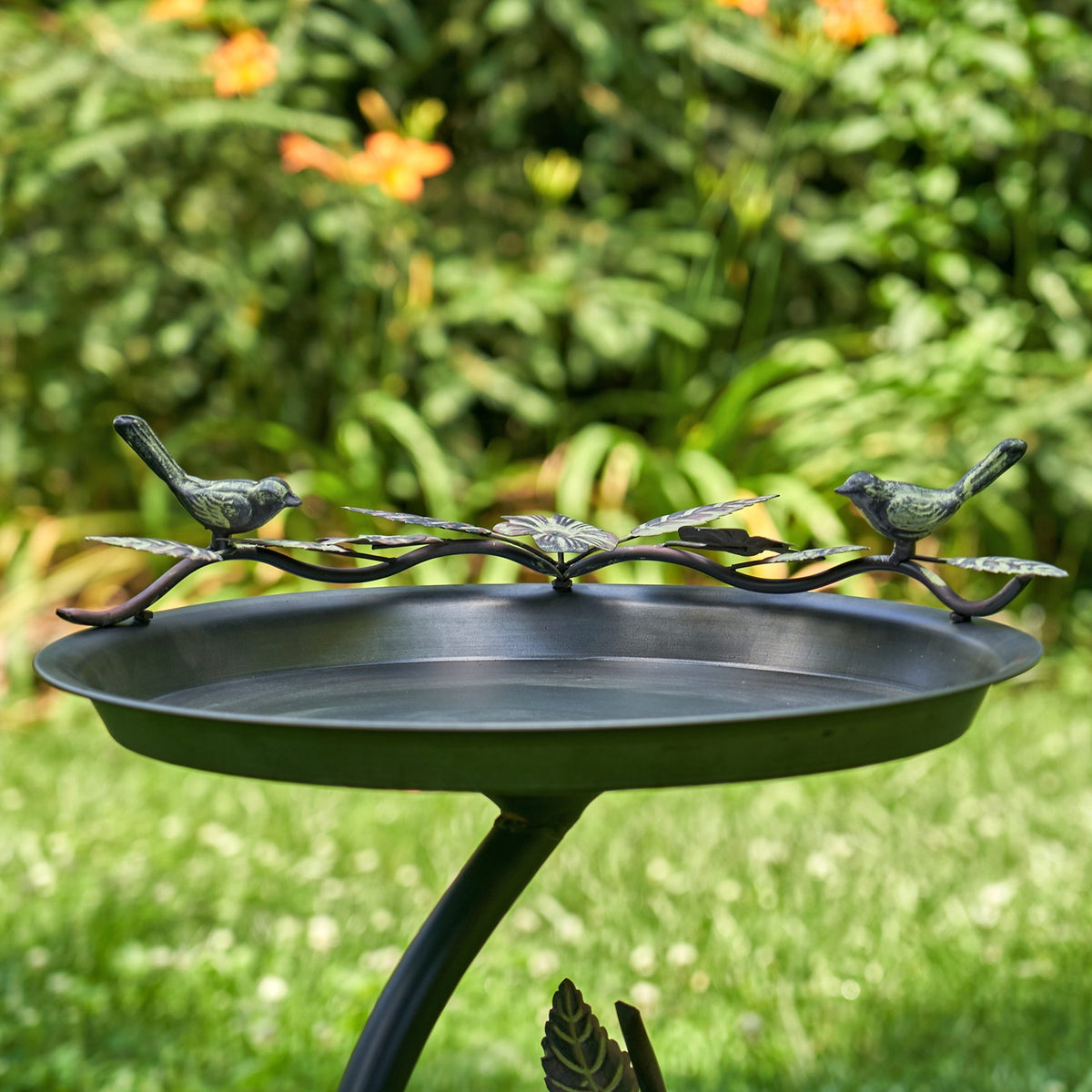 Shallow metal birdbath with a curved branch stand and three bird ornaments, set against a leafy garden.