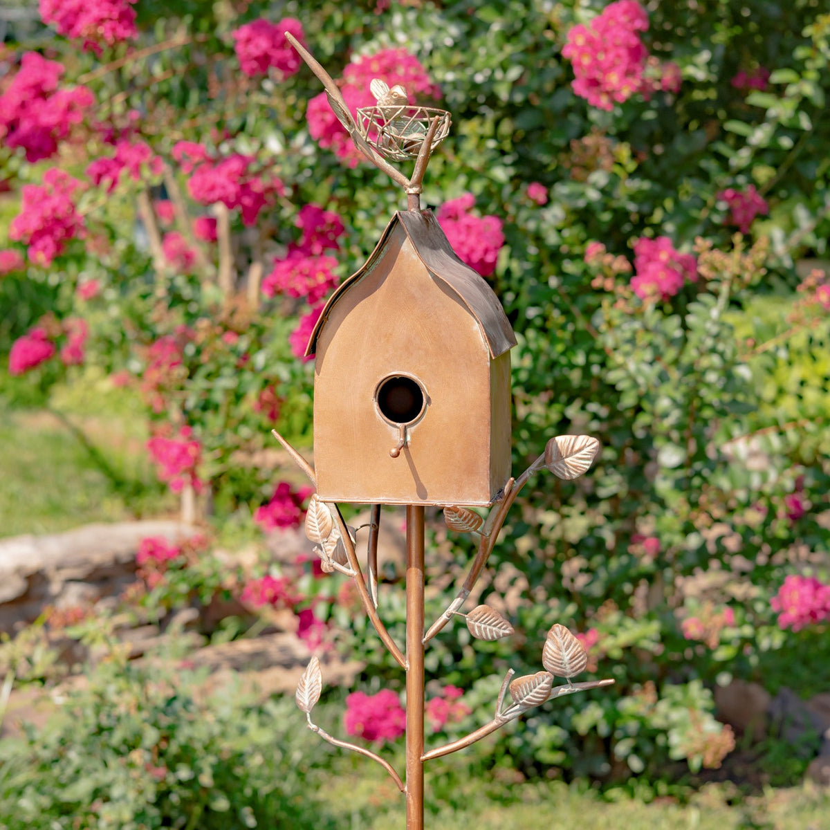 Front view of the birdhouse with its round entrance, sloped roof, leafy branches, and wire nest ornament.