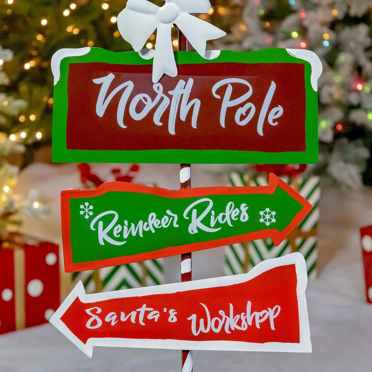 Close-up of three red-and-green directional signs on the striped pole, with wrapped gifts and snowy trees behind.