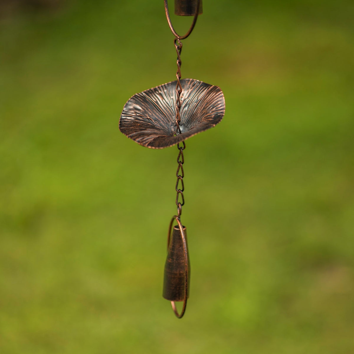 A fluted mushroom-shaped cup and the chain links between two bronze chimes, shown against greenery.