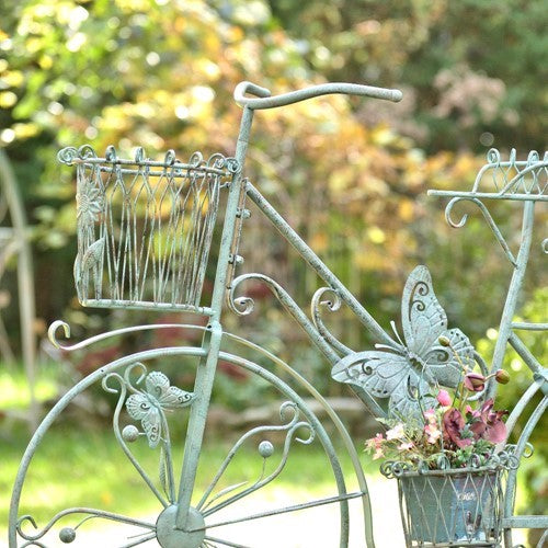 Close view of the bicycle stand’s handlebar, front basket, butterfly ornament, and flower-filled center basket.