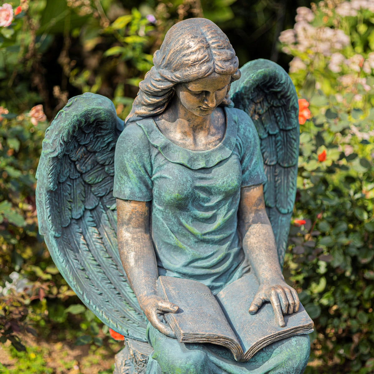 Seated angel statue looking down at an open book, with feathered wings and a weathered teal finish.