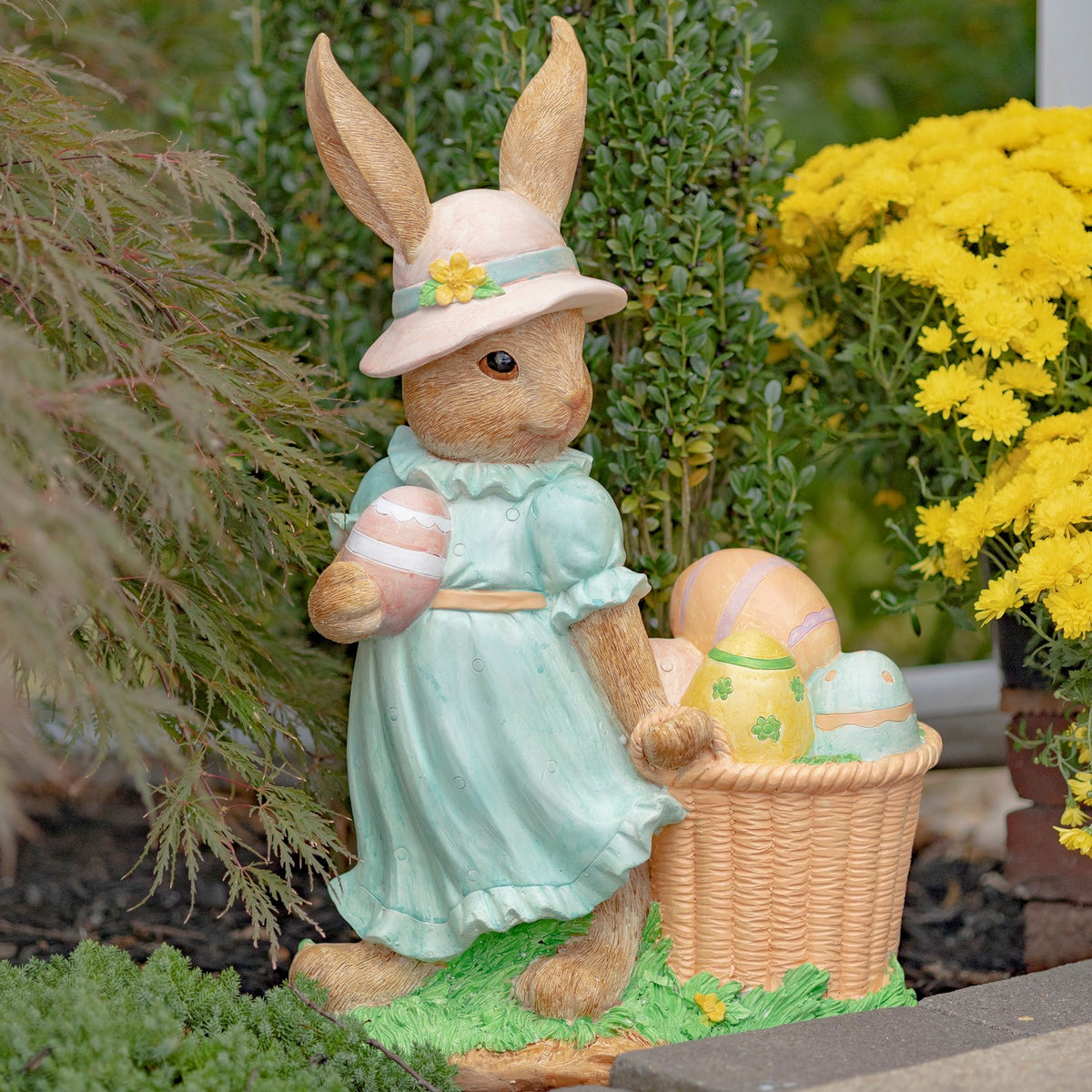 Rabbit statue in a blue dress poses beside an Easter egg basket among garden plants.
