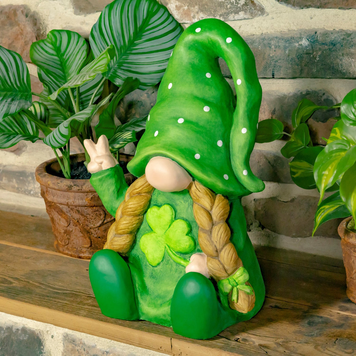 Seated green gnome with blond braids, a four-leaf clover on its chest, and one hand raised.