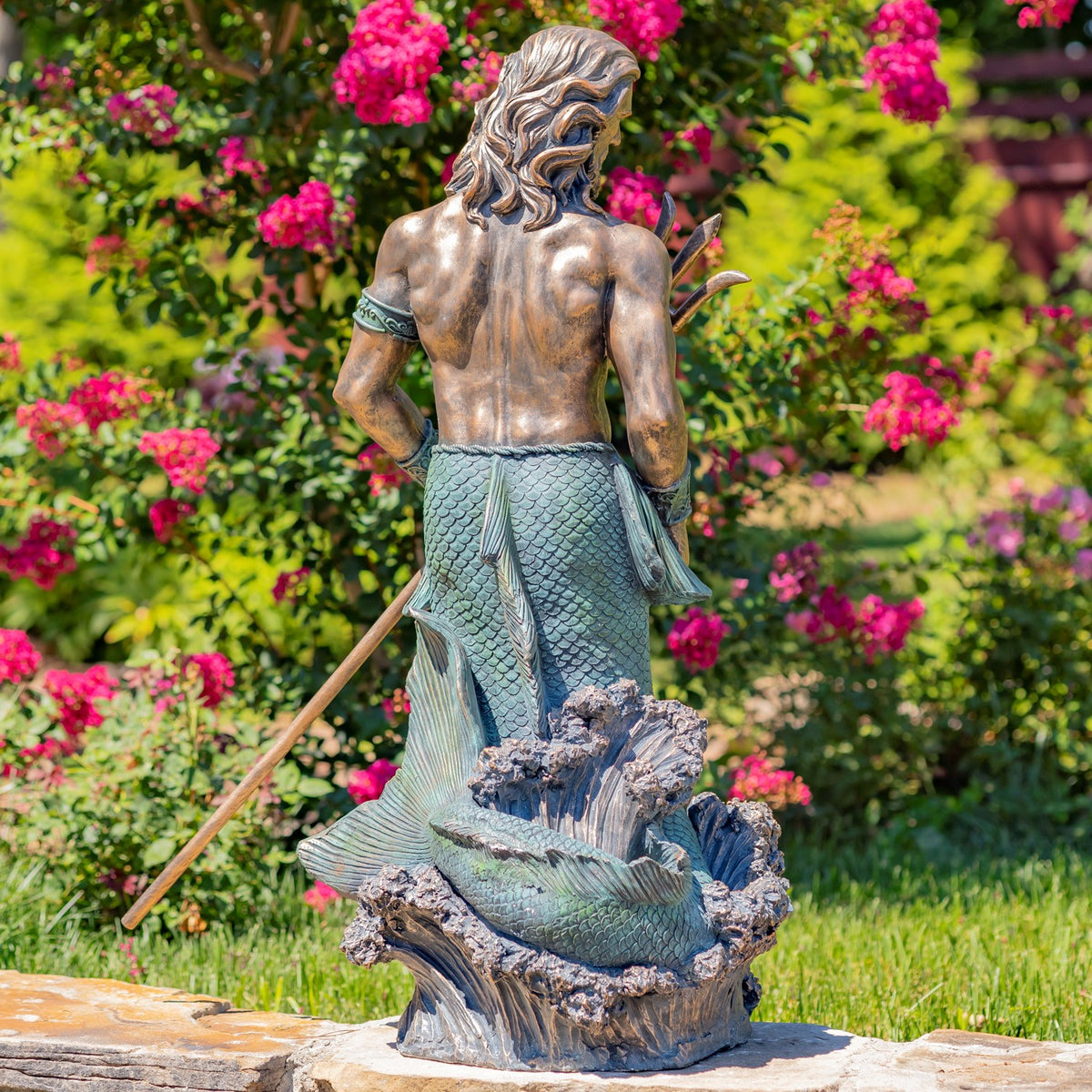 Back view of the merman statue, showing his long hair, scaled tail, and wave base in the garden.