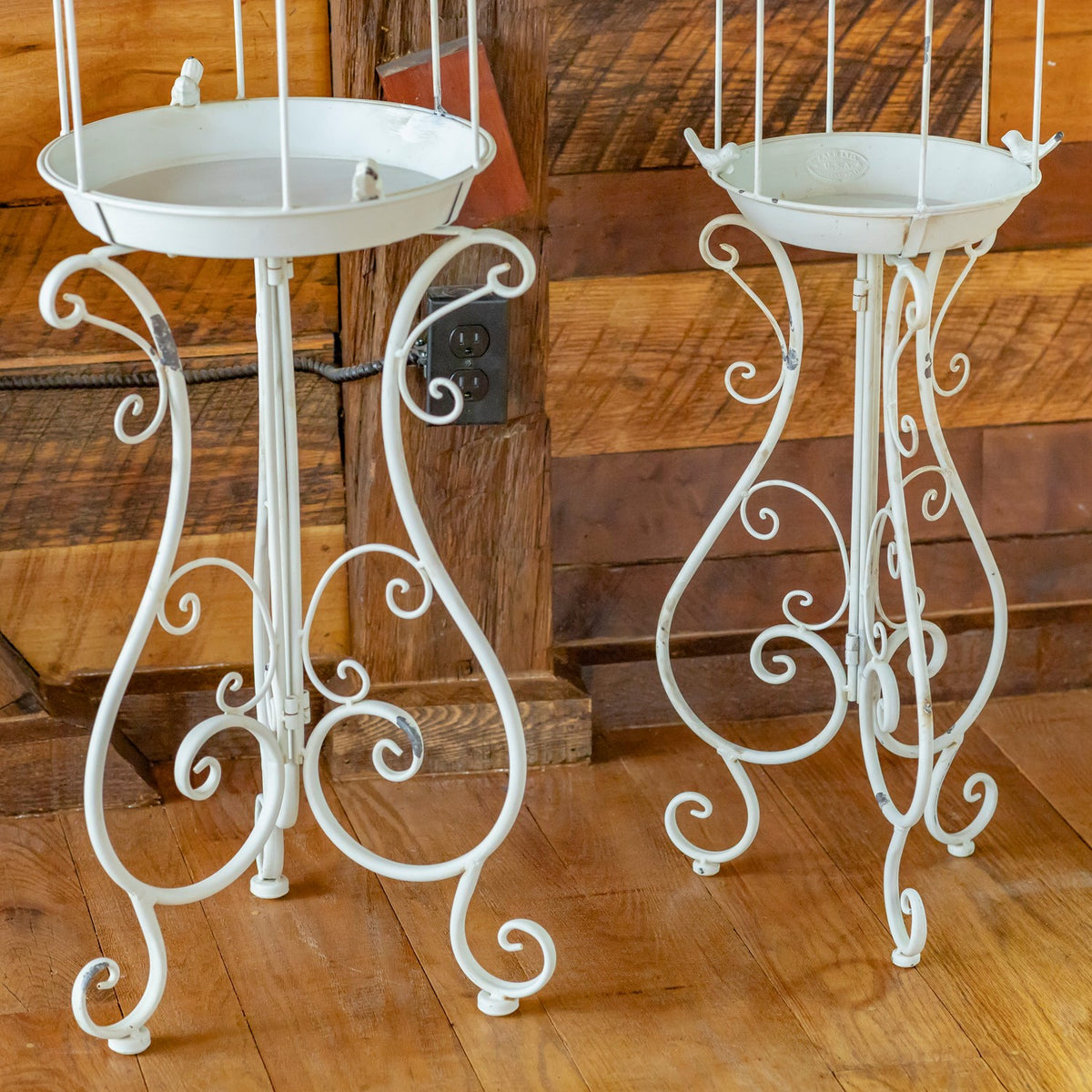 Two white iron stands with shallow round trays and curling scrollwork legs against a wood-paneled wall.
