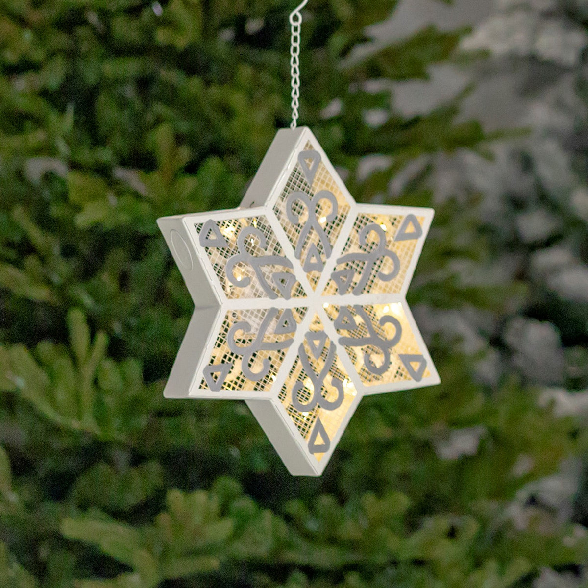 Angled view of a glowing white and silver snowflake hanging before evergreen branches