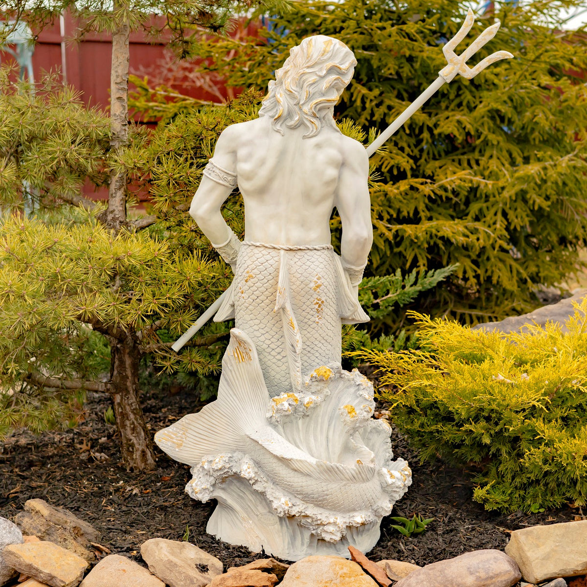 Back of the merman statue shows his hair, shoulders, scaled tail, and trident against garden shrubs.