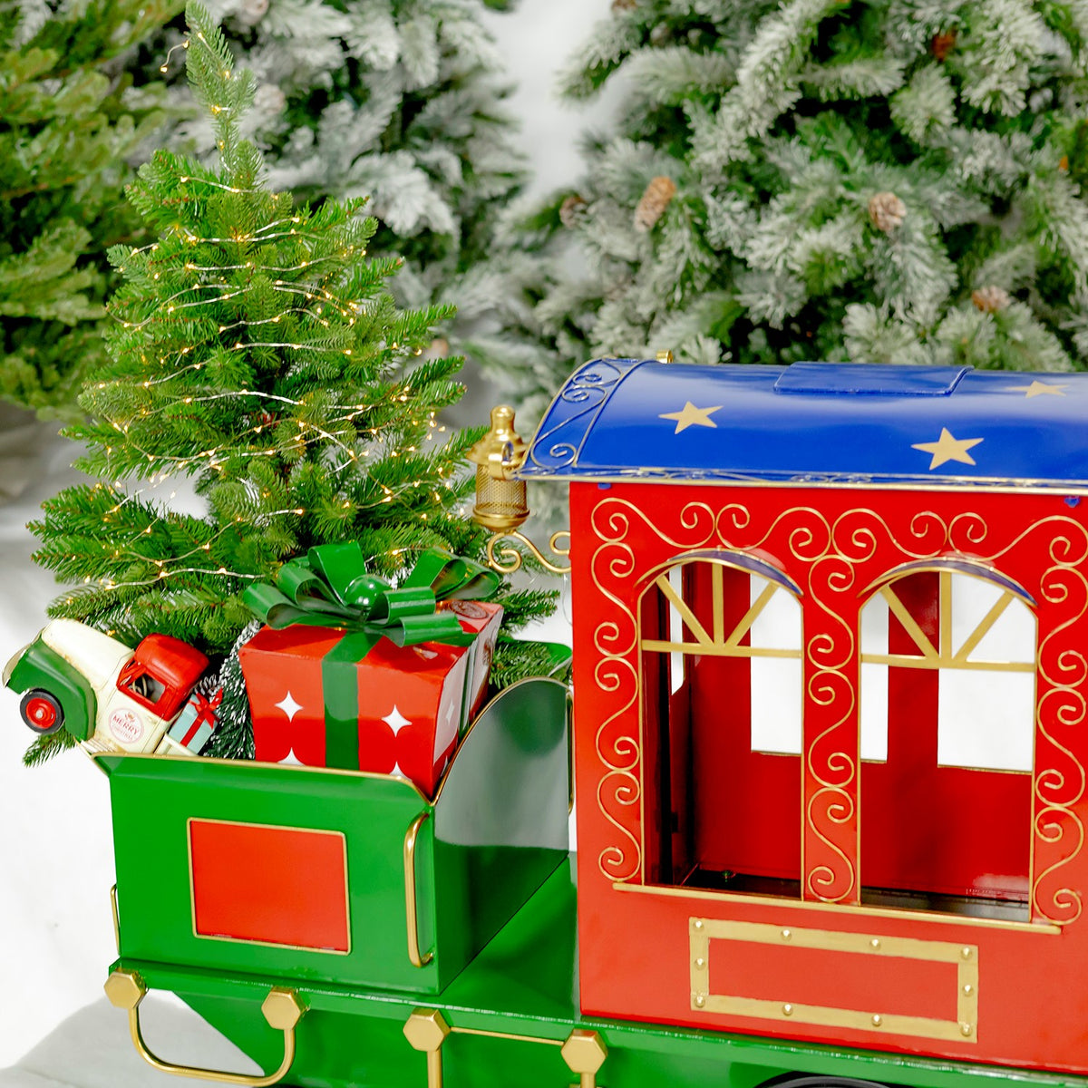 The train’s green rear cart holds wrapped gifts beside the red carriage, with a lit Christmas tree in the background.