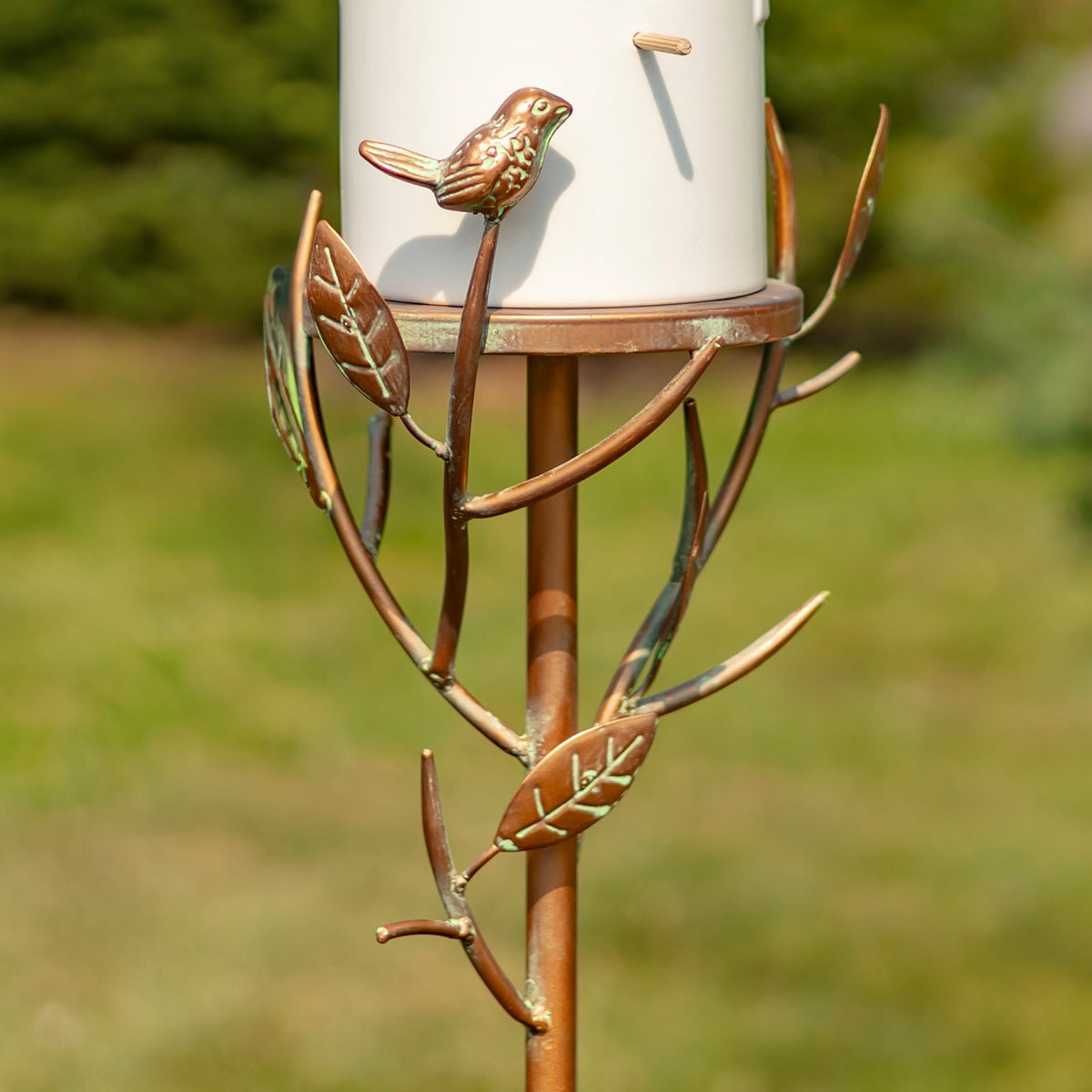 Metal branches and leaf shapes curl around the stake below the birdhouse, with a small bird ornament perched above.
