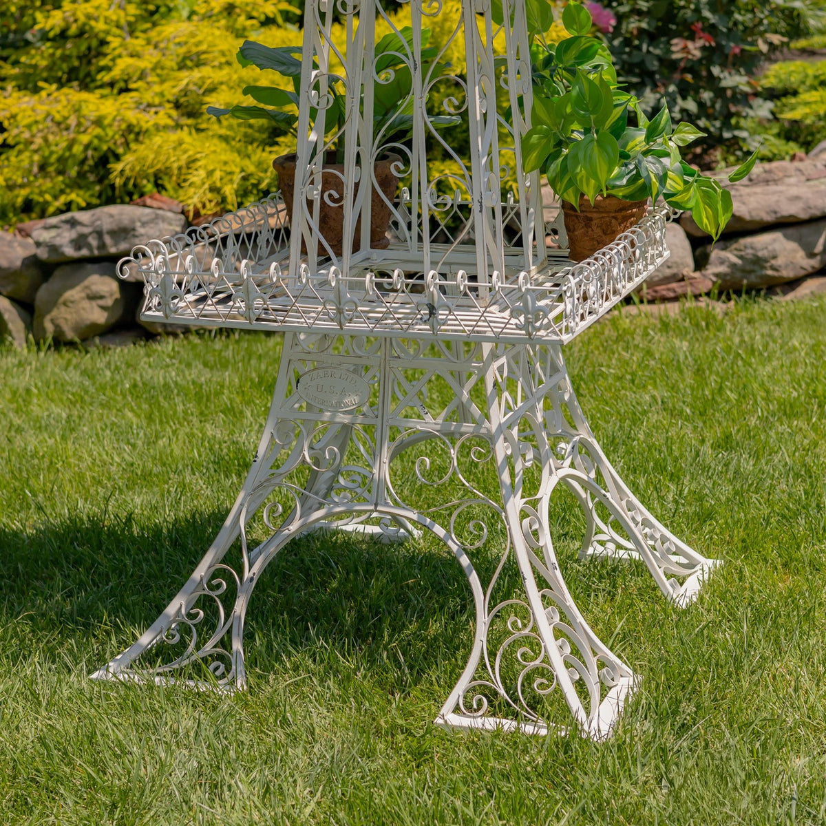 White scrollwork iron plant stand with a raised square tier and potted greenery on a lawn.