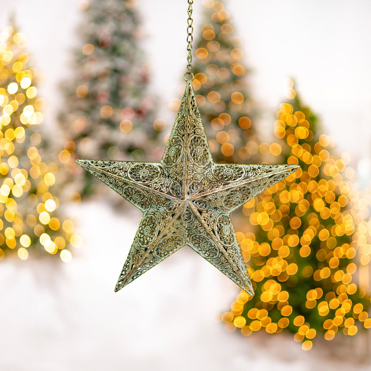 A patterned three-dimensional gold star hangs from a chain, with glowing Christmas lights behind it.